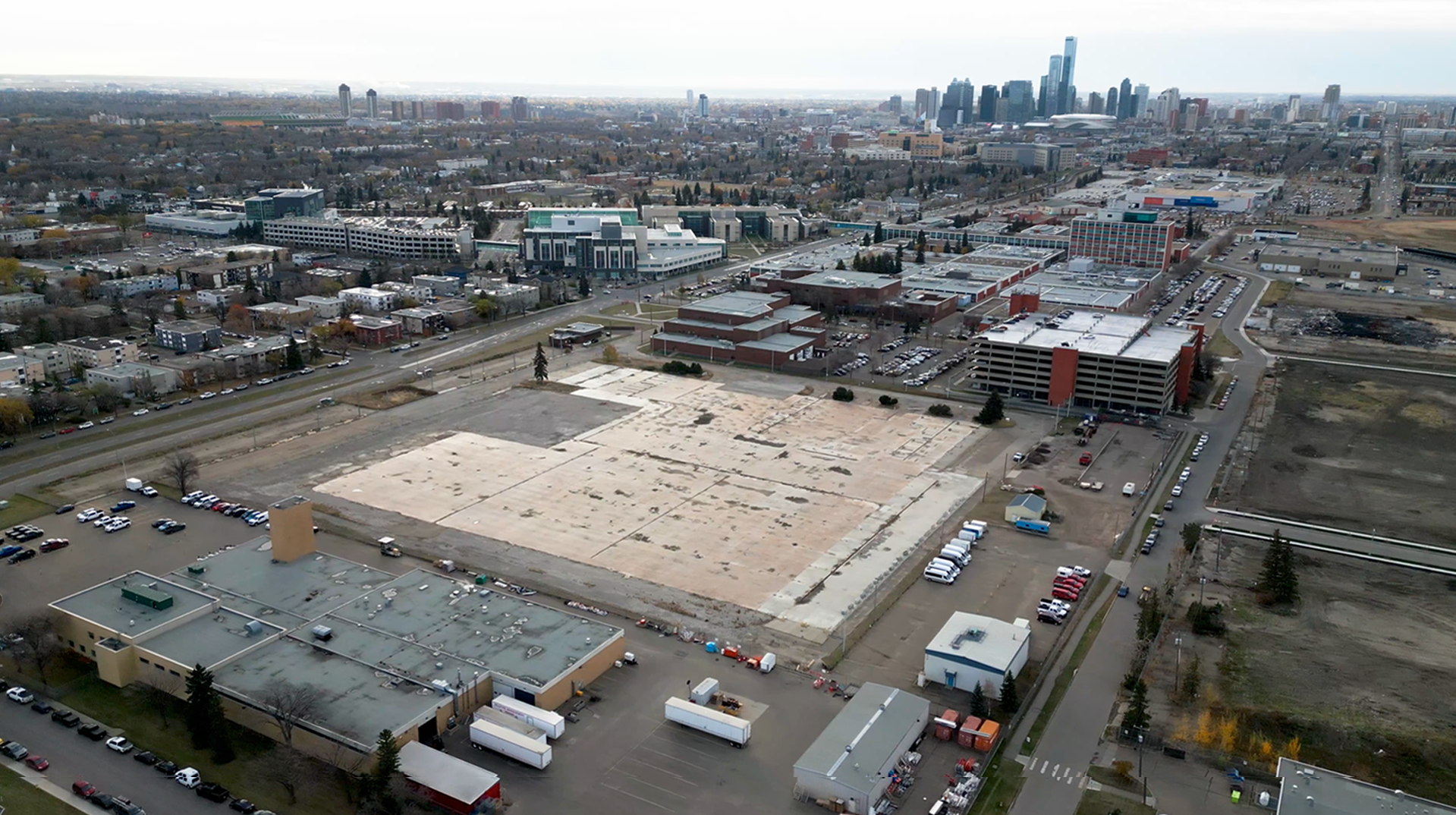 Aerial view of a large cleared construction site on a campus, surrounded by academic buildings, parking areas, and streets, with Edmonton’s downtown skyline visible in the distance under an overcast sky.