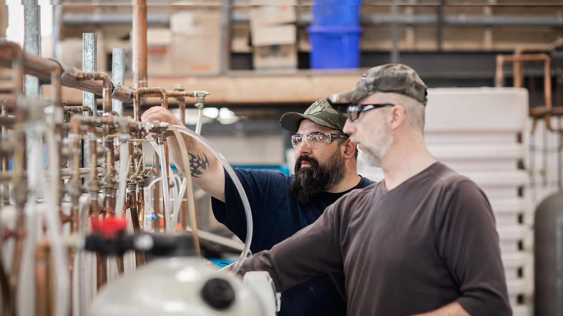 Two males wearing safety glasses stand at a workstation with copper piping. Two males wearing safety glasses stand at a workstation with copper piping.