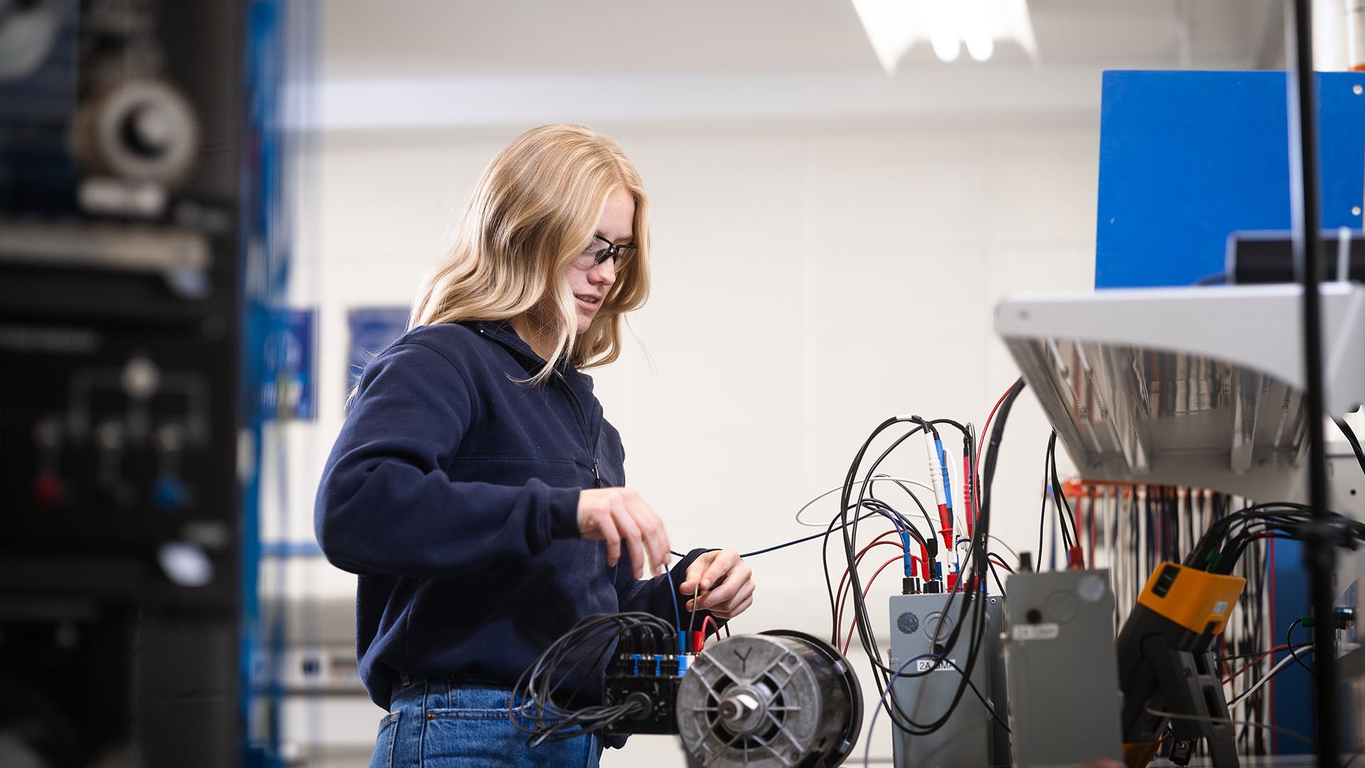 Female electrical student wearing safety glasses sits at a desk with electrical wires and electrician tools.