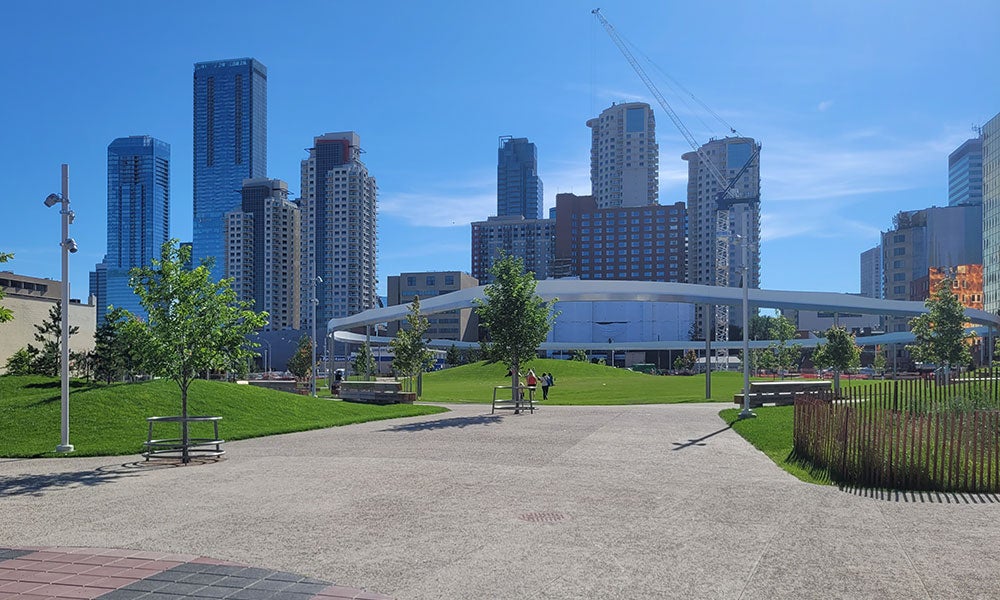 Landscaped urban park with wide pedestrian pathways, grassy lawns, benches, and a circular elevated feature, set against a backdrop of downtown high-rise buildings under a clear blue sky.