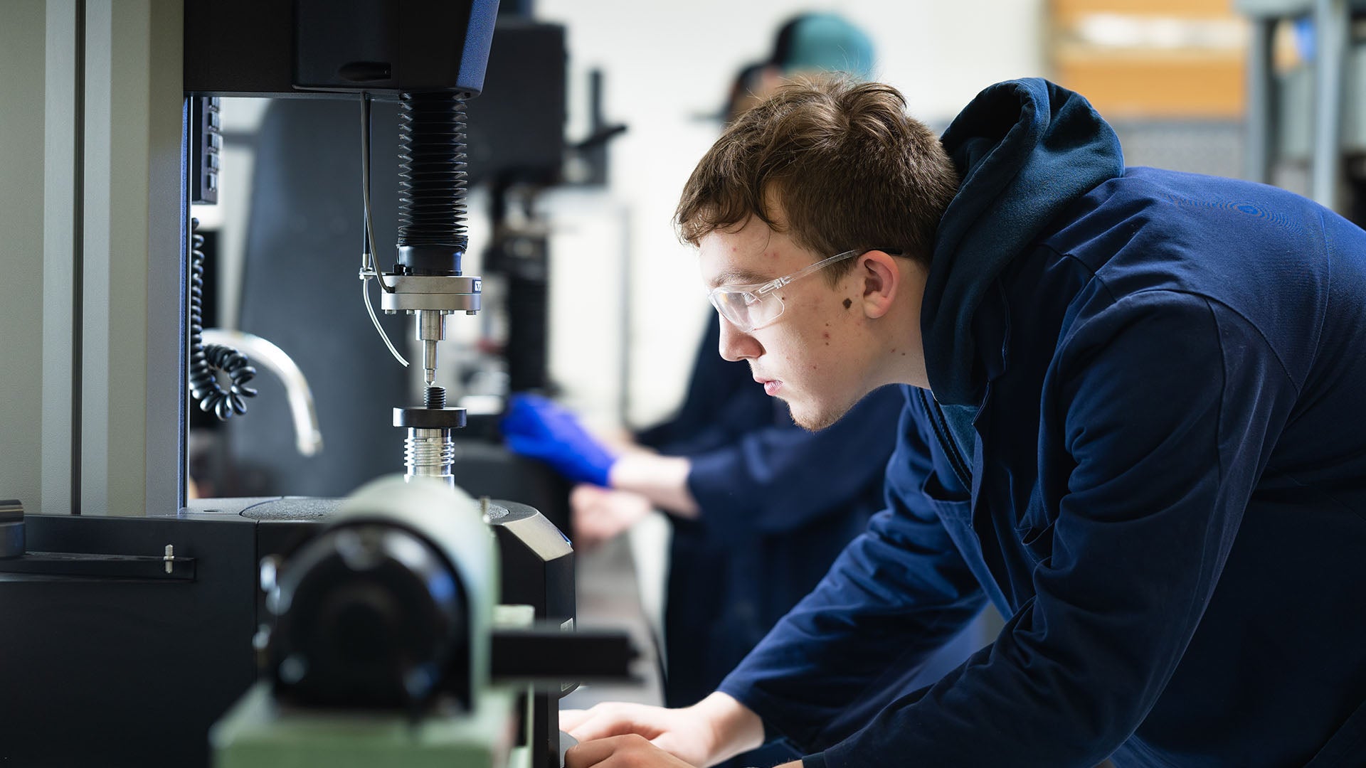 Male student with brown hair and safety glasses looks closely at a machine pressing down on material.