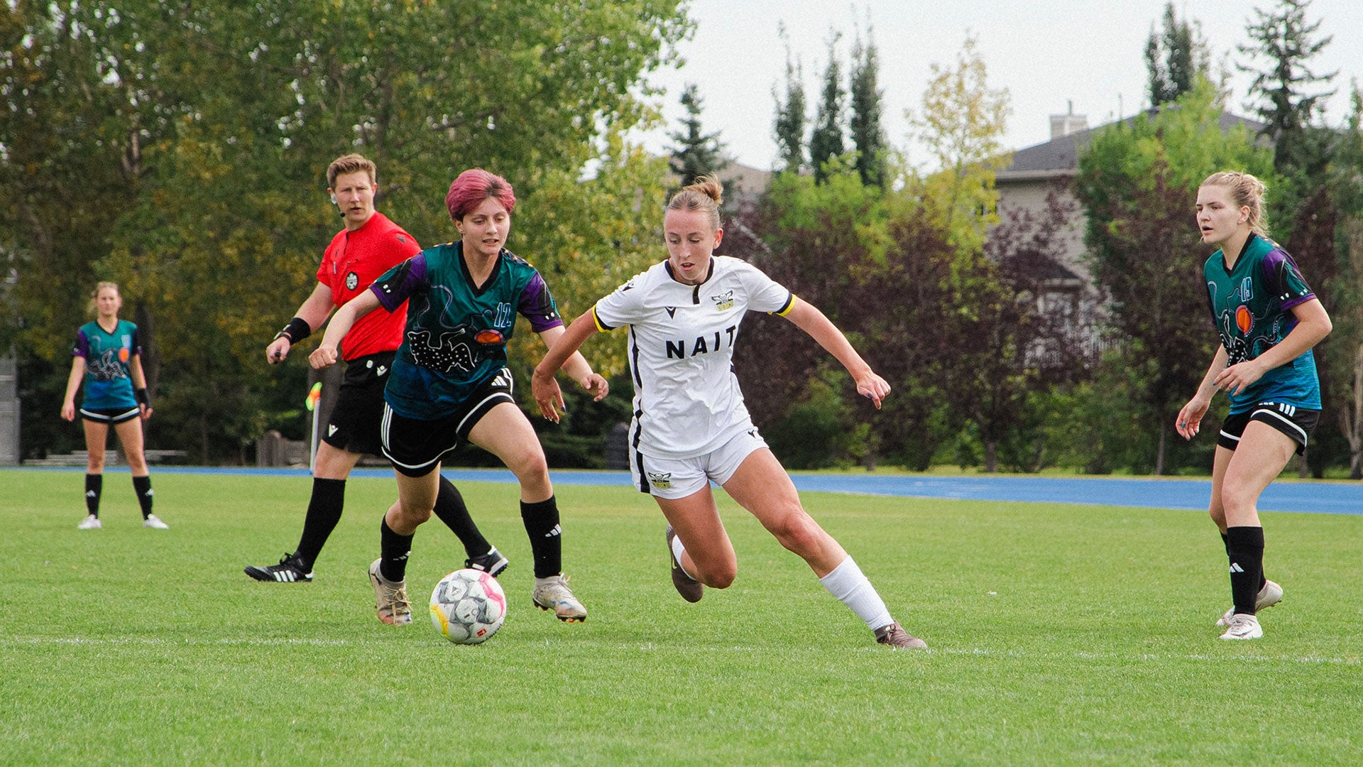 Women's soccer match in progress on a grassy field, with players competing for the ball and a referee observing play in an outdoor athletic setting. Women's soccer match in progress on a grassy field, with players competing for the ball and a referee observing play in an outdoor athletic setting.