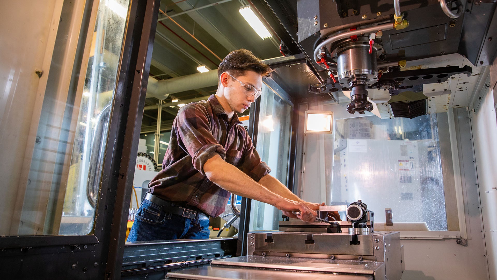 A machinist student secures a metal piece inside of a CNC milling machine. A machinist student secures a metal piece inside of a CNC milling machine.