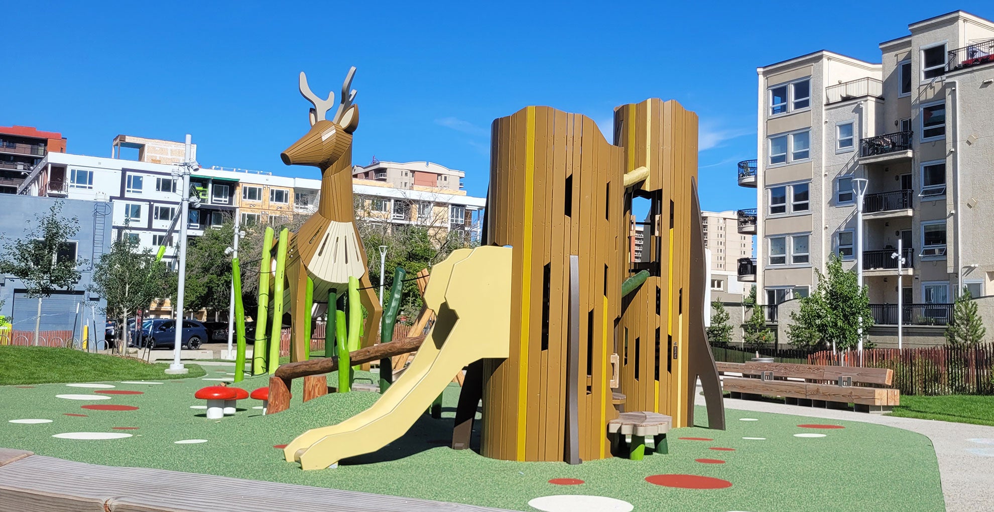 Outdoor playground featuring a deer-themed climbing structure with a slide on a rubberized play surface, surrounded by modern apartment buildings under a clear blue sky. Outdoor playground featuring a deer-themed climbing structure with a slide on a rubberized play surface, surrounded by modern apartment buildings under a clear blue sky.