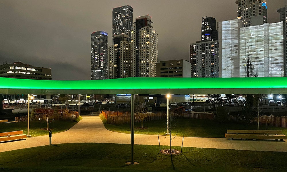 Nighttime view of a landscaped urban park with illuminated pathways and a glowing green elevated feature, set against a backdrop of high-rise buildings and city lights.