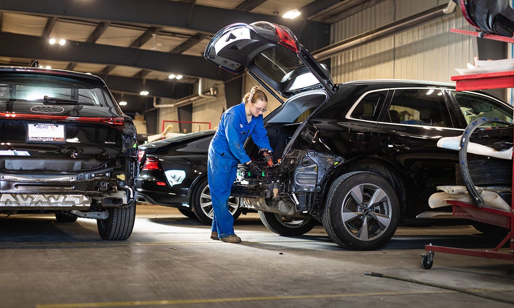 Person in blue coveralls working on an open vehicle in a busy automotive repair shop with multiple cars and service equipment visible.