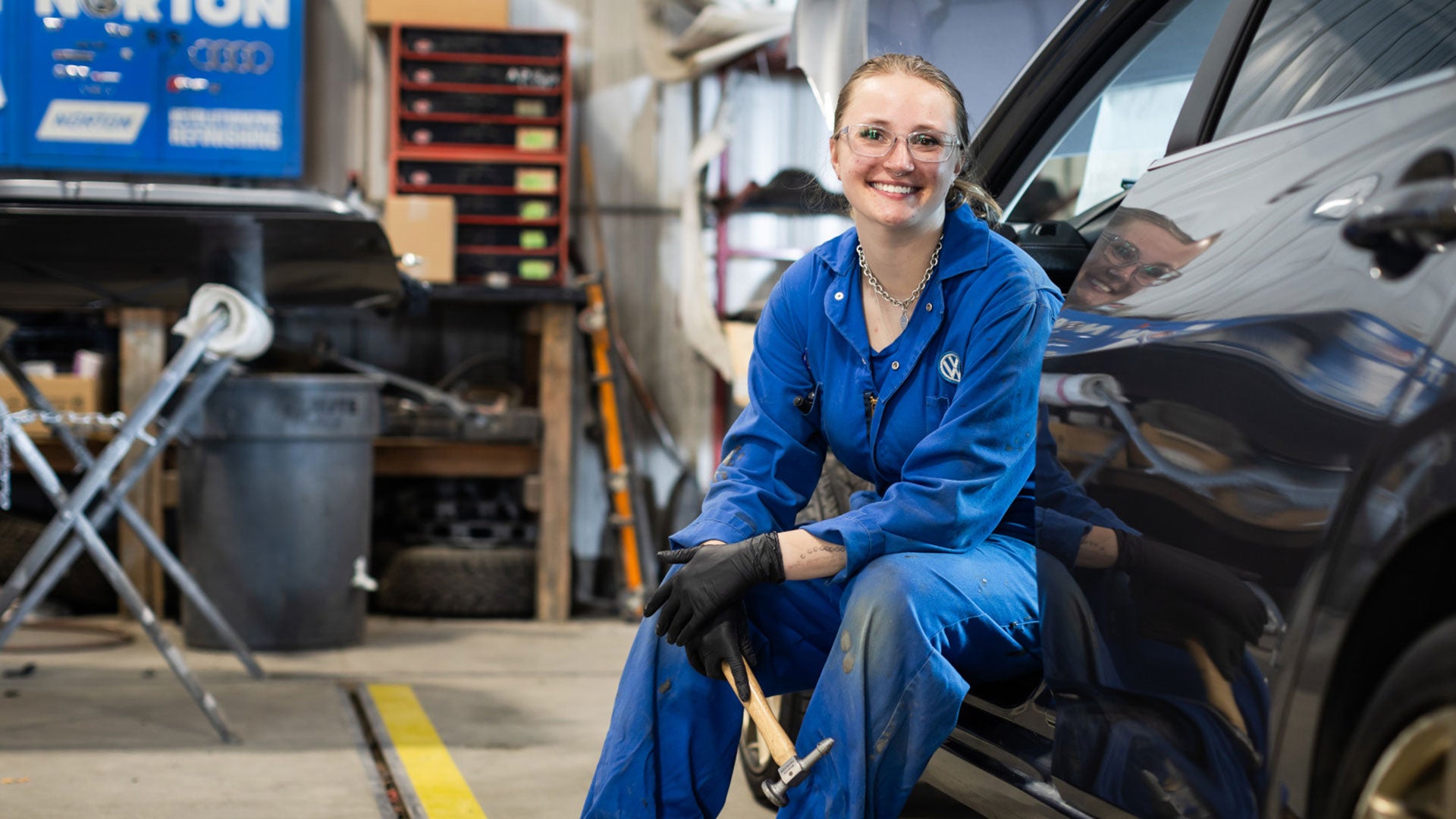 Person in blue coveralls kneeling beside a vehicle in an auto repair shop, with tools and equipment visible in the background. Person in blue coveralls kneeling beside a vehicle in an auto repair shop, with tools and equipment visible in the background.