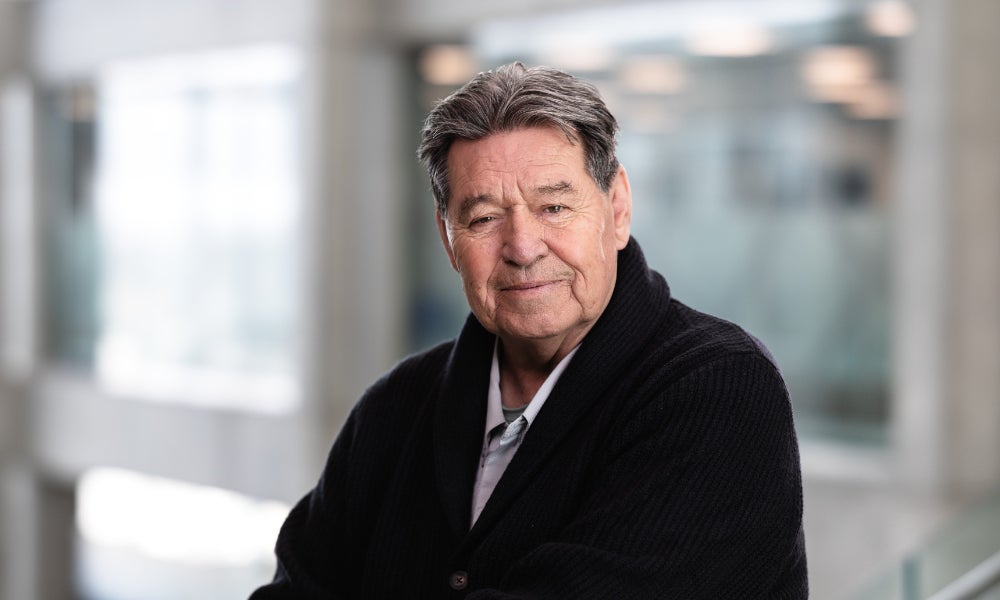 Head-and-shoulders portrait of a person with short gray hair wearing a dark sweater, standing indoors in a bright modern setting with a softly blurred background.