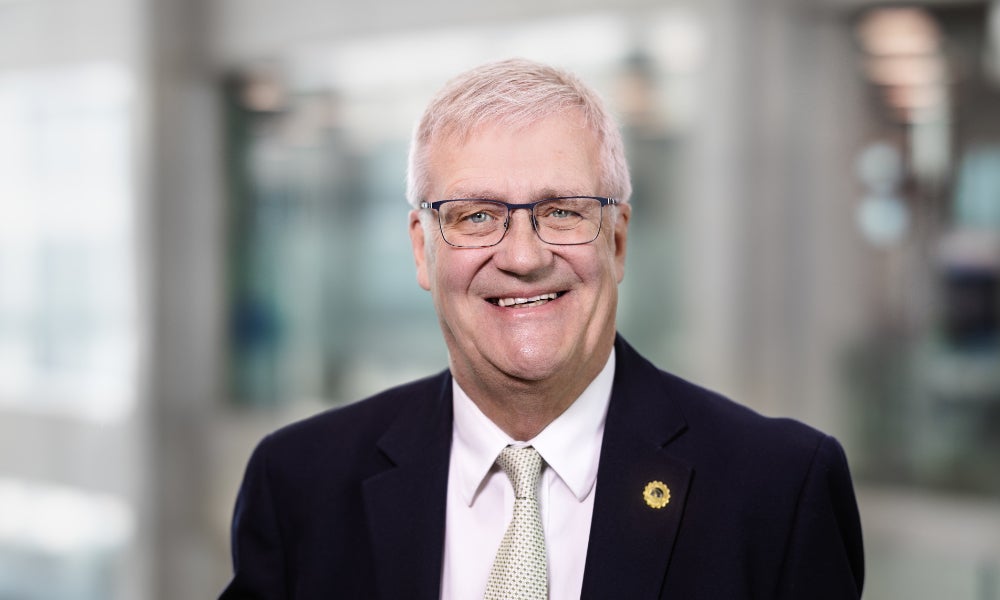 Head-and-shoulders portrait of a person with short light hair and glasses wearing a dark suit and light tie, standing indoors in a bright modern setting with a softly blurred background.