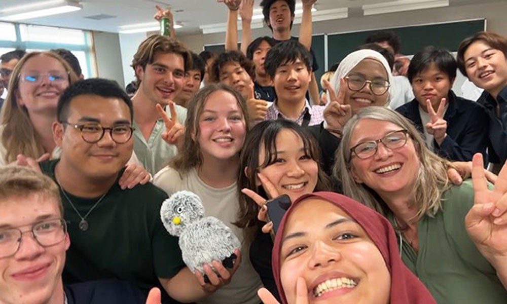 A large group of people crowded together indoors, posing closely for a group selfie while several hold up peace signs and one person in front holds a small gray plush toy.