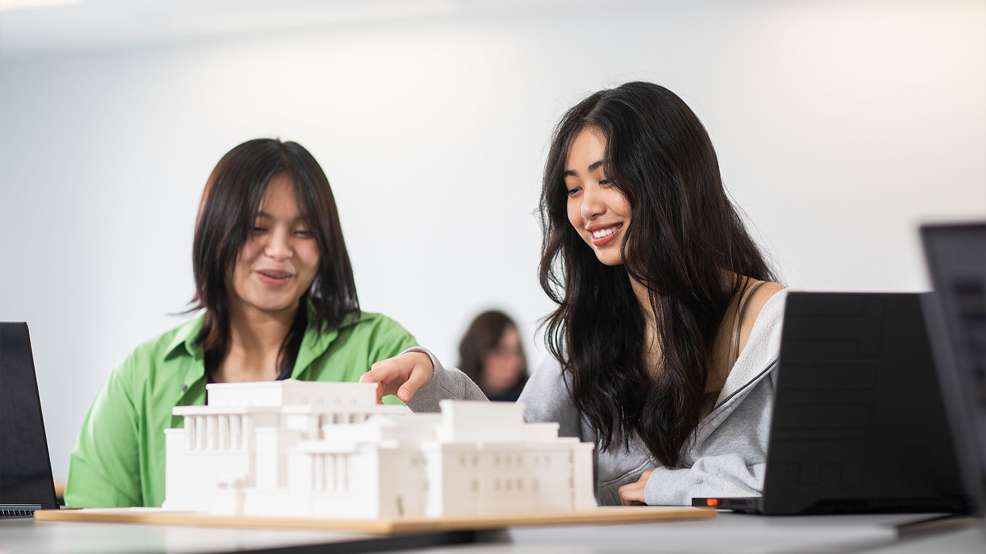 Two women collaborating on an architectural model, smiling while discussing, with a laptop open nearby.