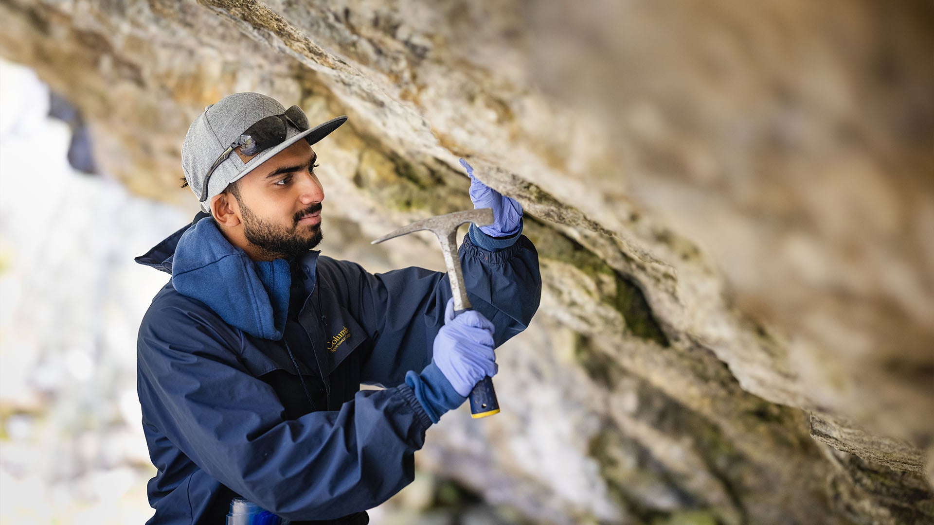 A geoscience technology student is at a rock site, using a geological hammer to chisel pieces of rock for examination or sampling.