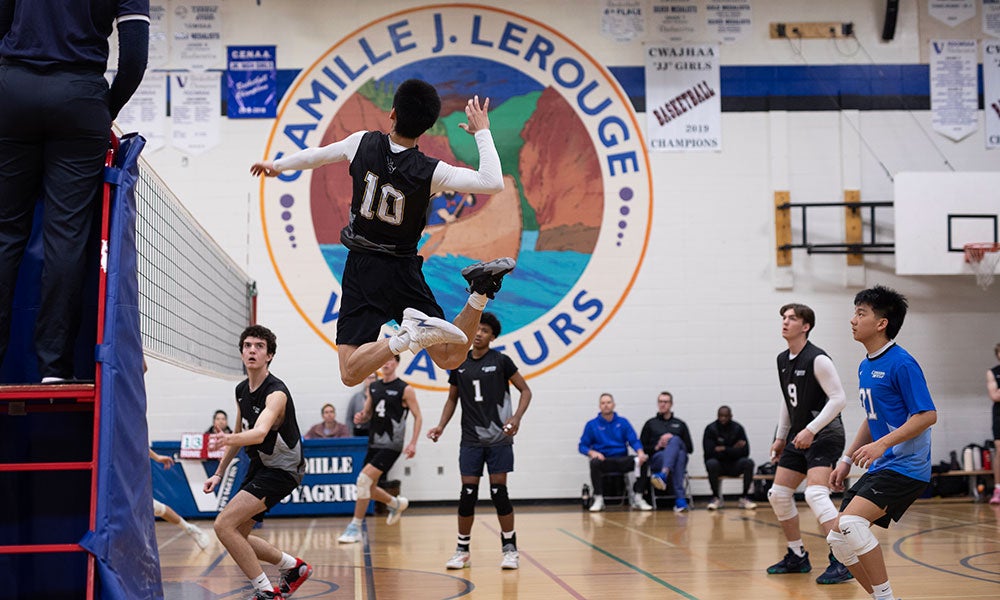 Players compete in an indoor volleyball match, with one player jumping near the net as teammates and opponents prepare for the play in a school gymnasium.