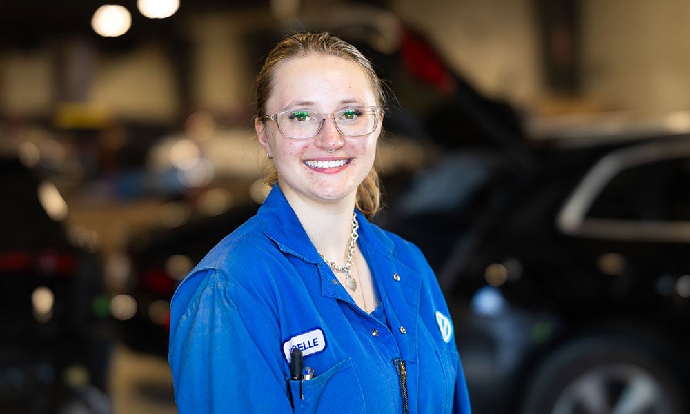 Close-up portrait of a person in blue coveralls standing in an automotive repair shop, with vehicles and workshop equipment blurred in the background.
