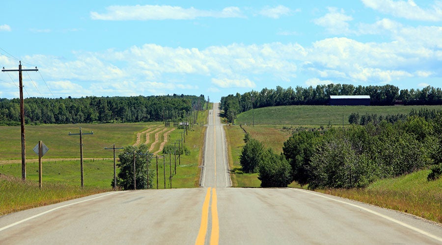 Two-lane rural road stretching through rolling countryside, bordered by fields, trees, utility poles, and a distant farm building under a blue sky with scattered clouds.