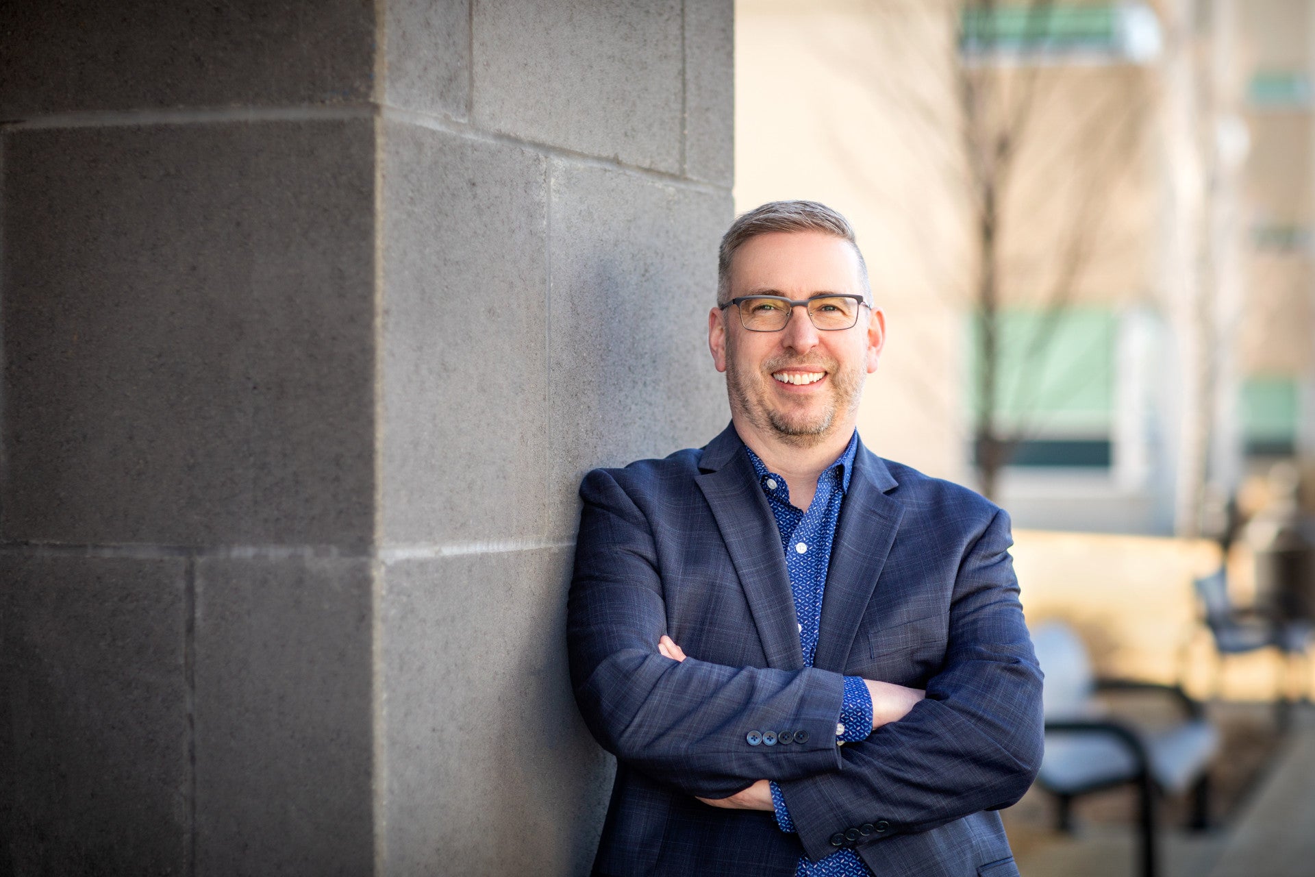 Professional portrait of a senior post-secondary leader standing with arms crossed beside a stone column on campus. The individual is dressed in business attire, with a blurred background featuring college buildings and outdoor seating areas.