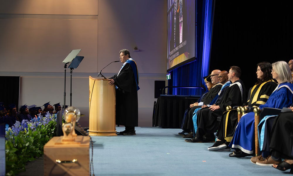 Speaker at podium addressing a graduation ceremony, with seated faculty in academic robes on stage and a large screen behind.