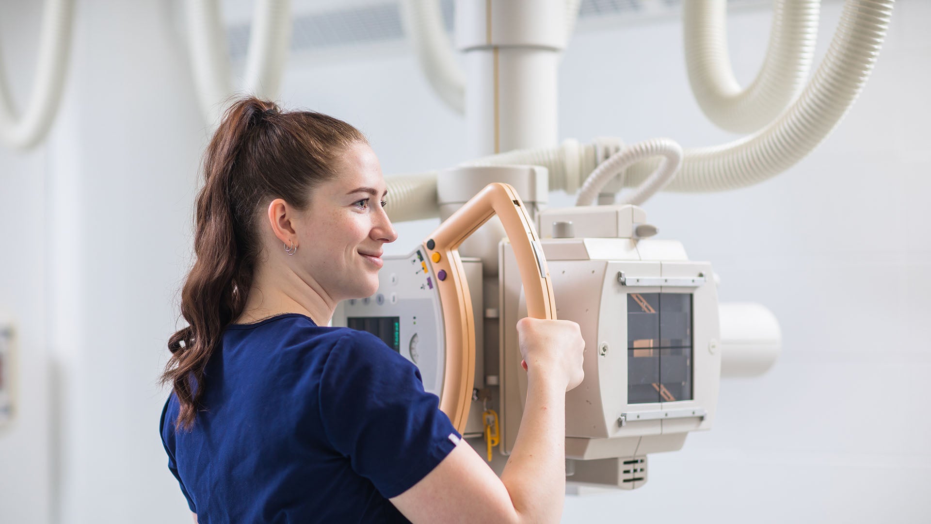 A medical radiologic technologist wearing blue scrubs is holding and adjusting the X-ray machine while smiling.