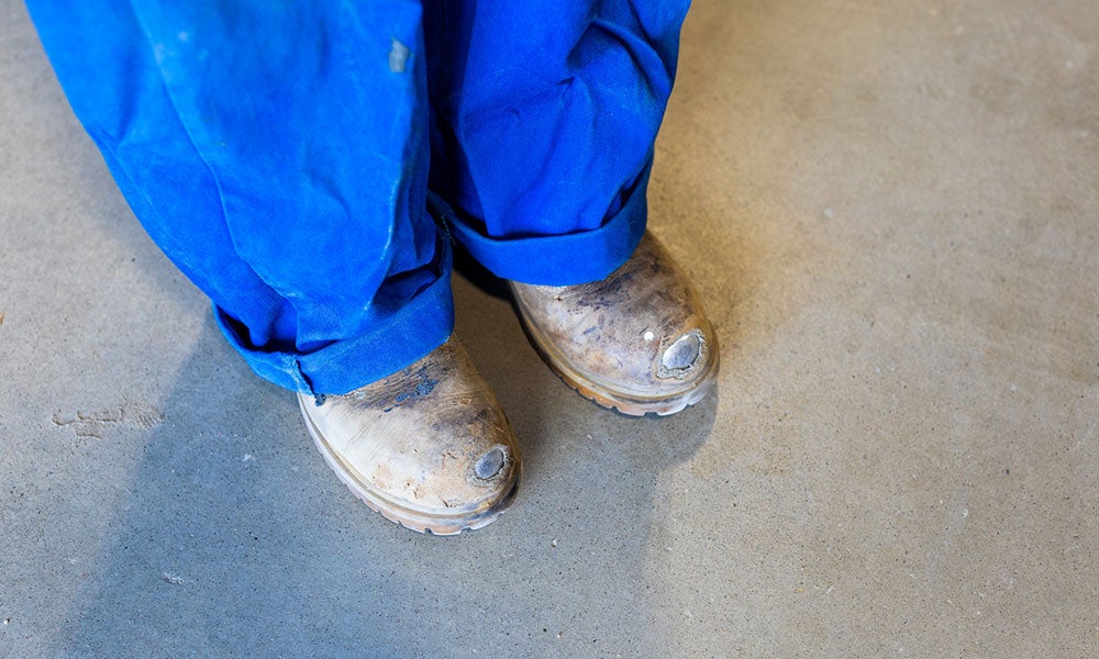 Close-up of worn work boots and blue coveralls standing on a concrete workshop floor.