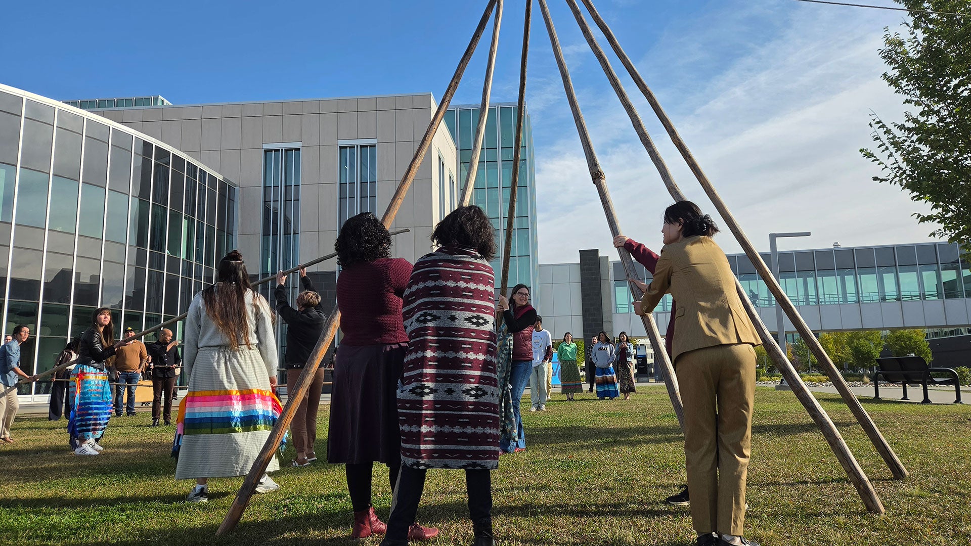 People raise a tipi framework on a grassy lawn outside a modern building while others watch and participate. People raise a tipi framework on a grassy lawn outside a modern building while others watch and participate.