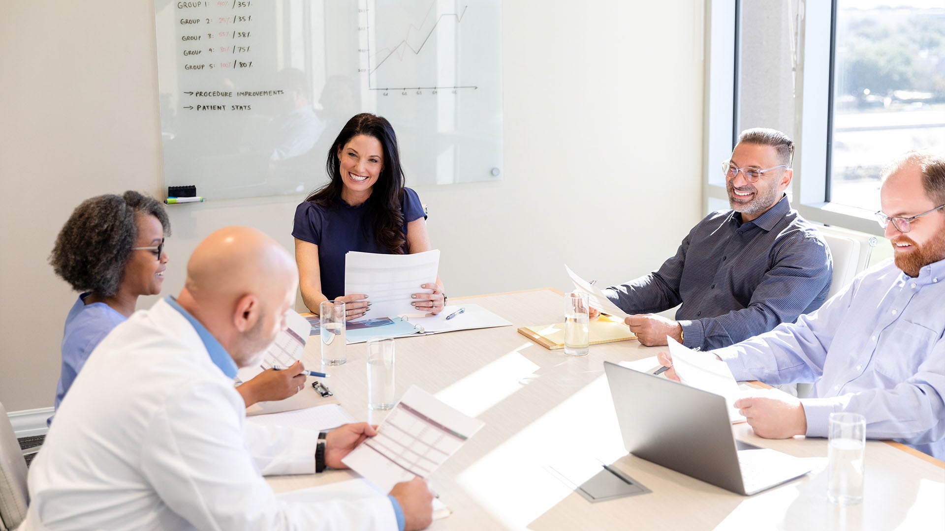 Smiling medical professionals sit at a bright table looking at documents and laptops.