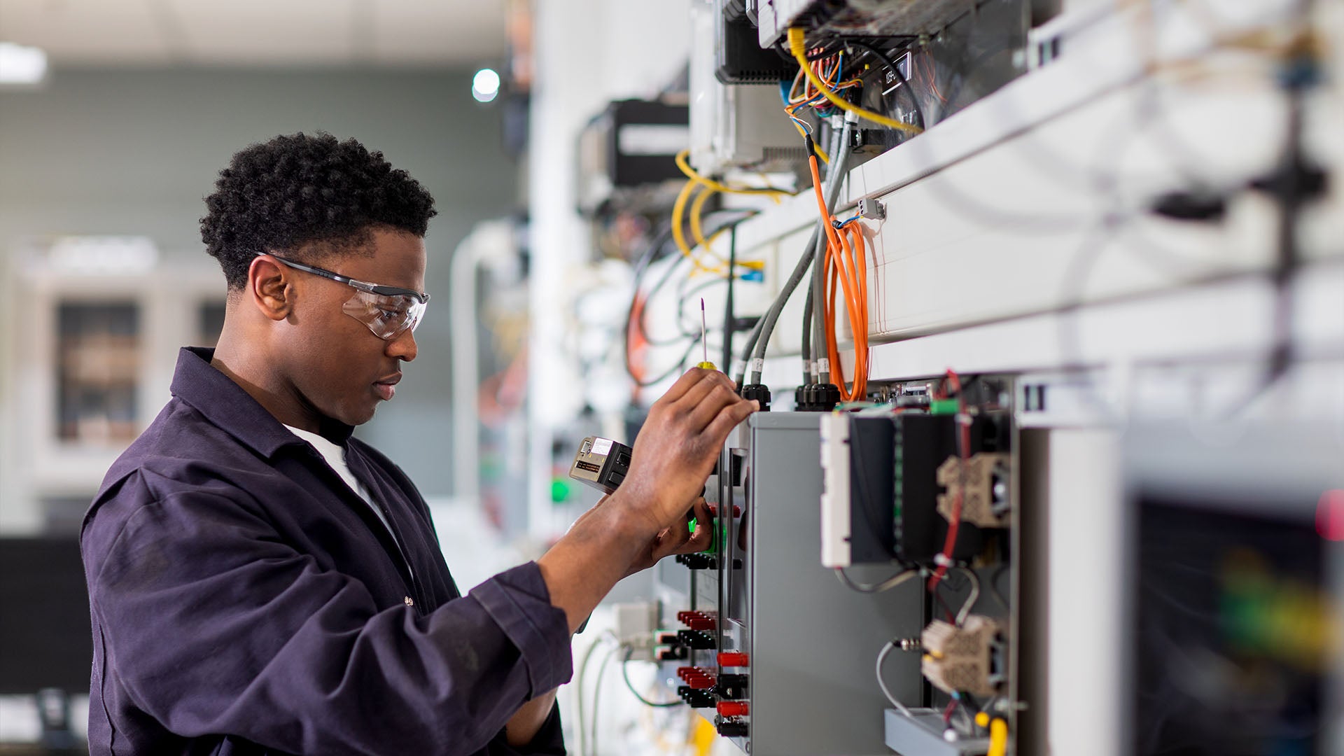 A man wearing safety glasses and a dark blue work jacket inspects an instrumentation panel.