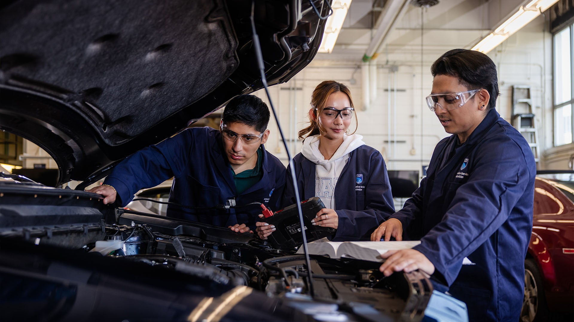 Three Automotive Service Technician students working under the hood of a vehicle. Three Automotive Service Technician students working under the hood of a vehicle.