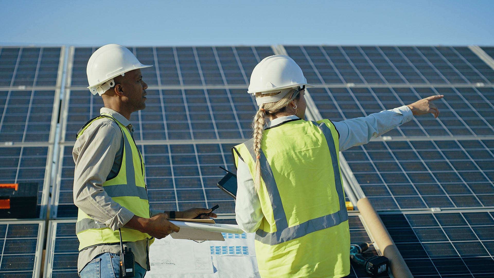 Two sustainability managers wearing hard hats and safety vests, holding documents, and equipped with walkie-talkies on-site. One is pointing in a direction while the other is looking at it.