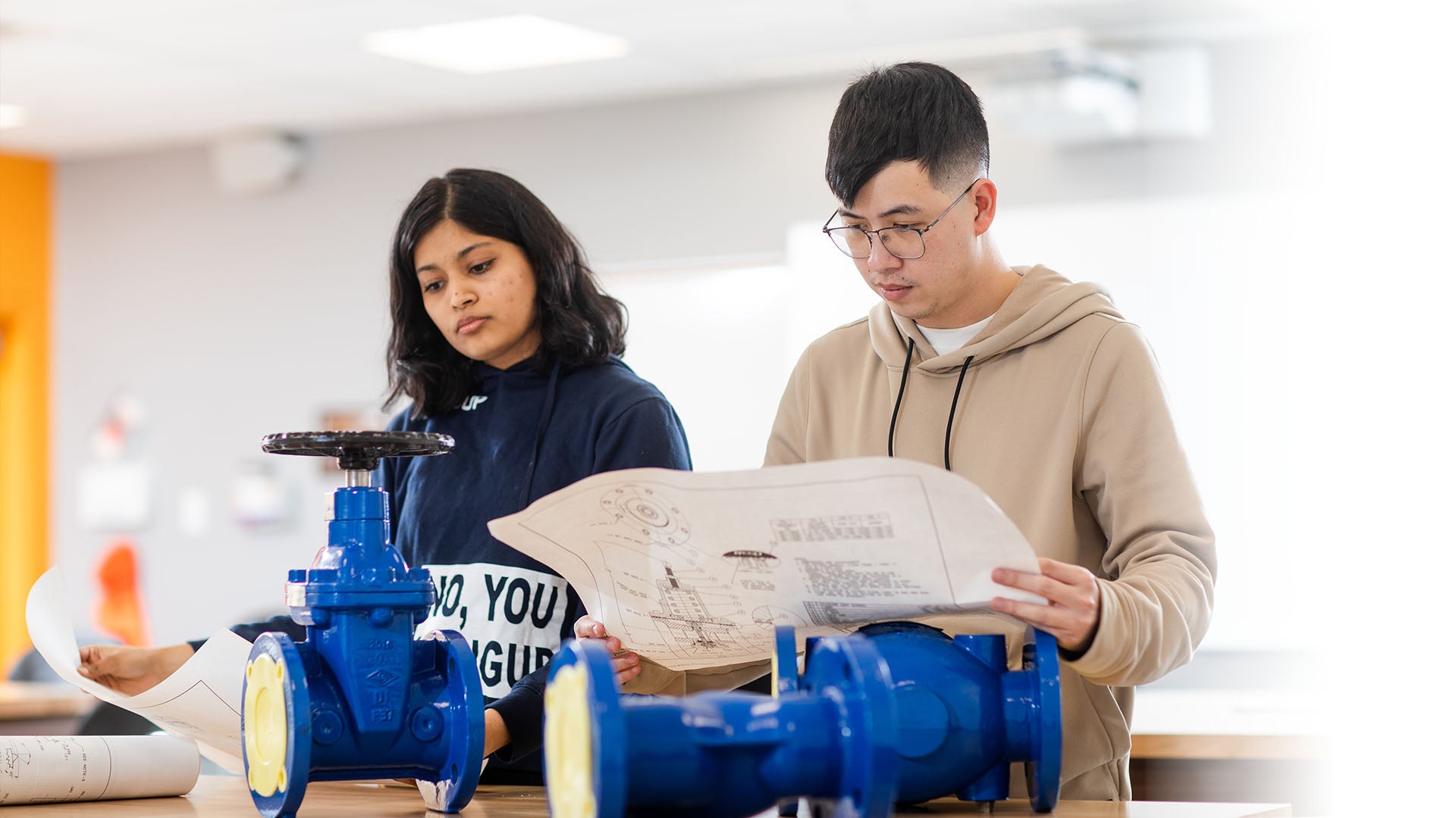 Two people working with blue industrial valves, examining technical blueprints in a classroom setting.