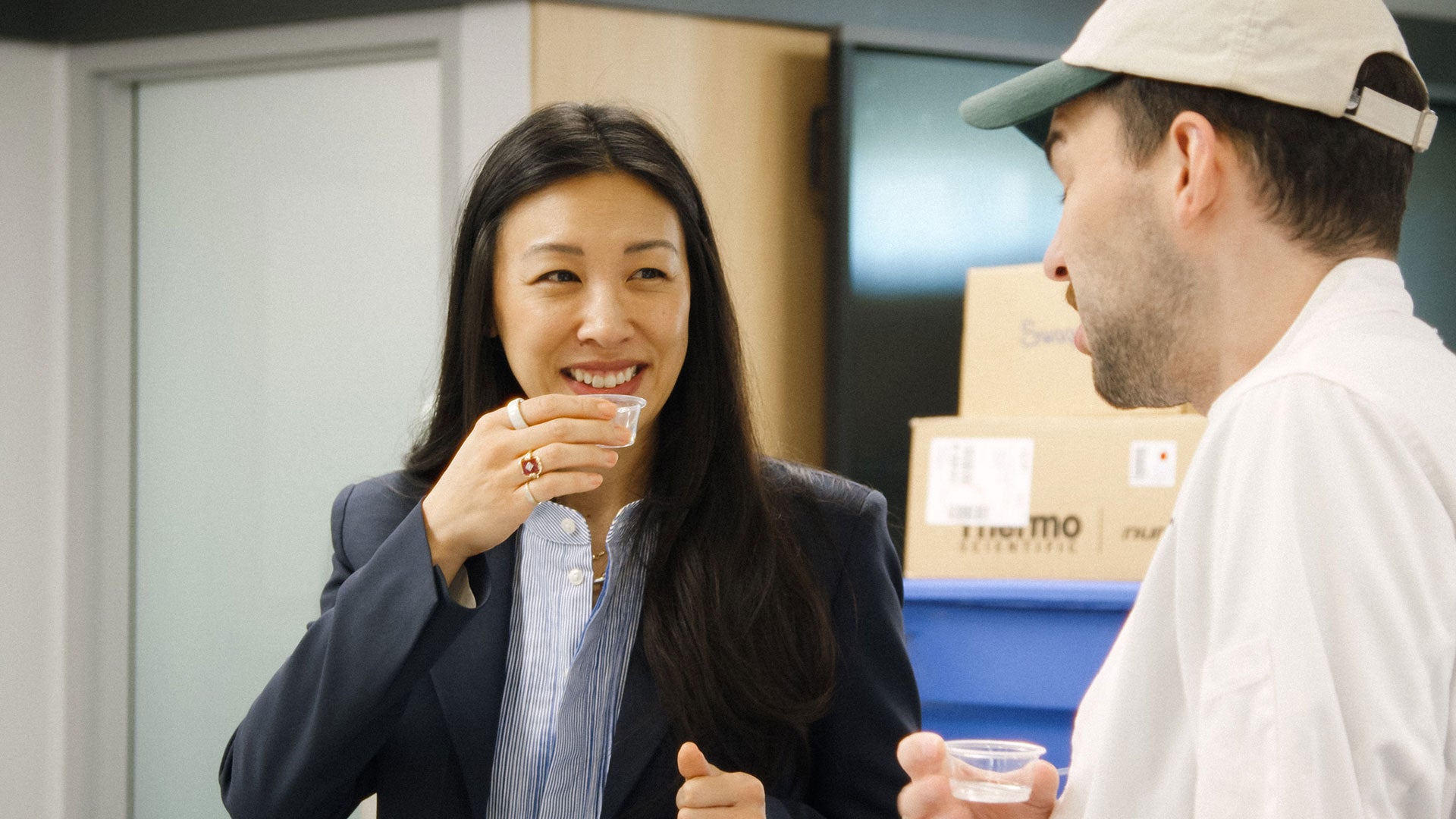 Person in business attire and person wearing a cap share a conversation while holding small cups in an office setting with stacked boxes in the background. Person in business attire and person wearing a cap share a conversation while holding small cups in an office setting with stacked boxes in the background.
