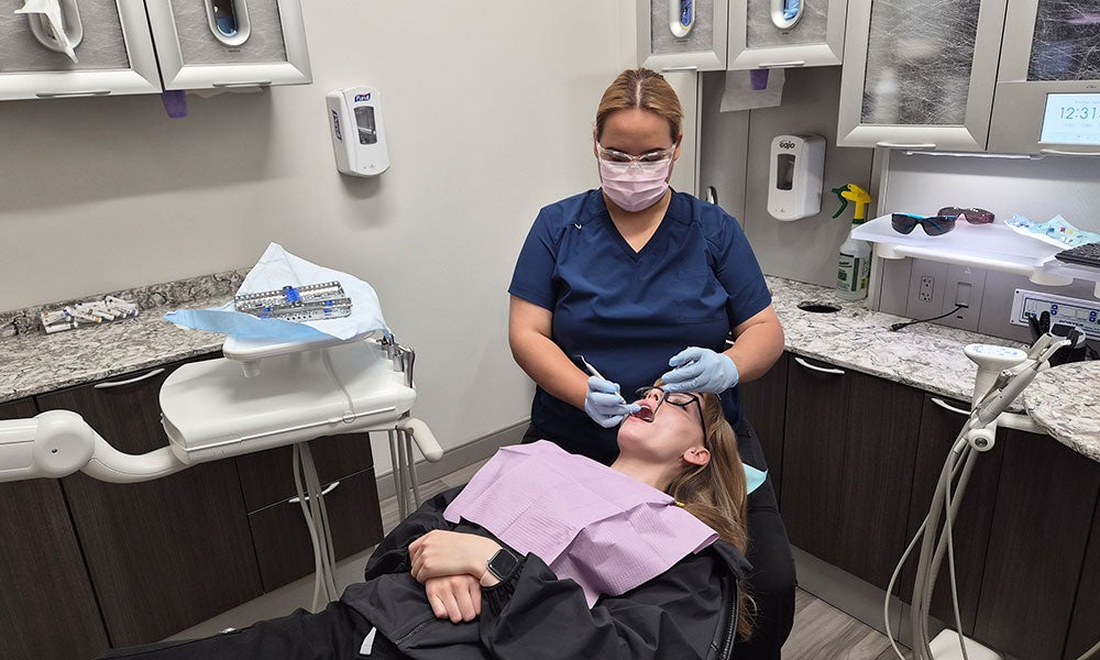 Person in blue scrubs and protective gear performing a dental procedure on a patient lying in a clinic chair inside a medical exam room.