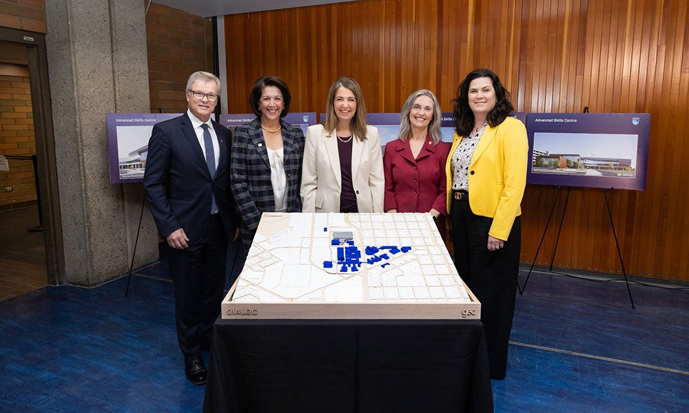 Five people, including Alberta Premier Danielle Smith, stand behind a campus development model display, with project renderings shown on easels in the background.