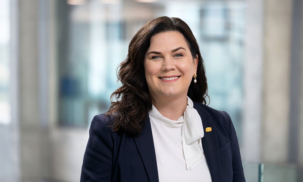 Professional headshot of a person wearing a navy blazer and white blouse, standing in a bright office setting.