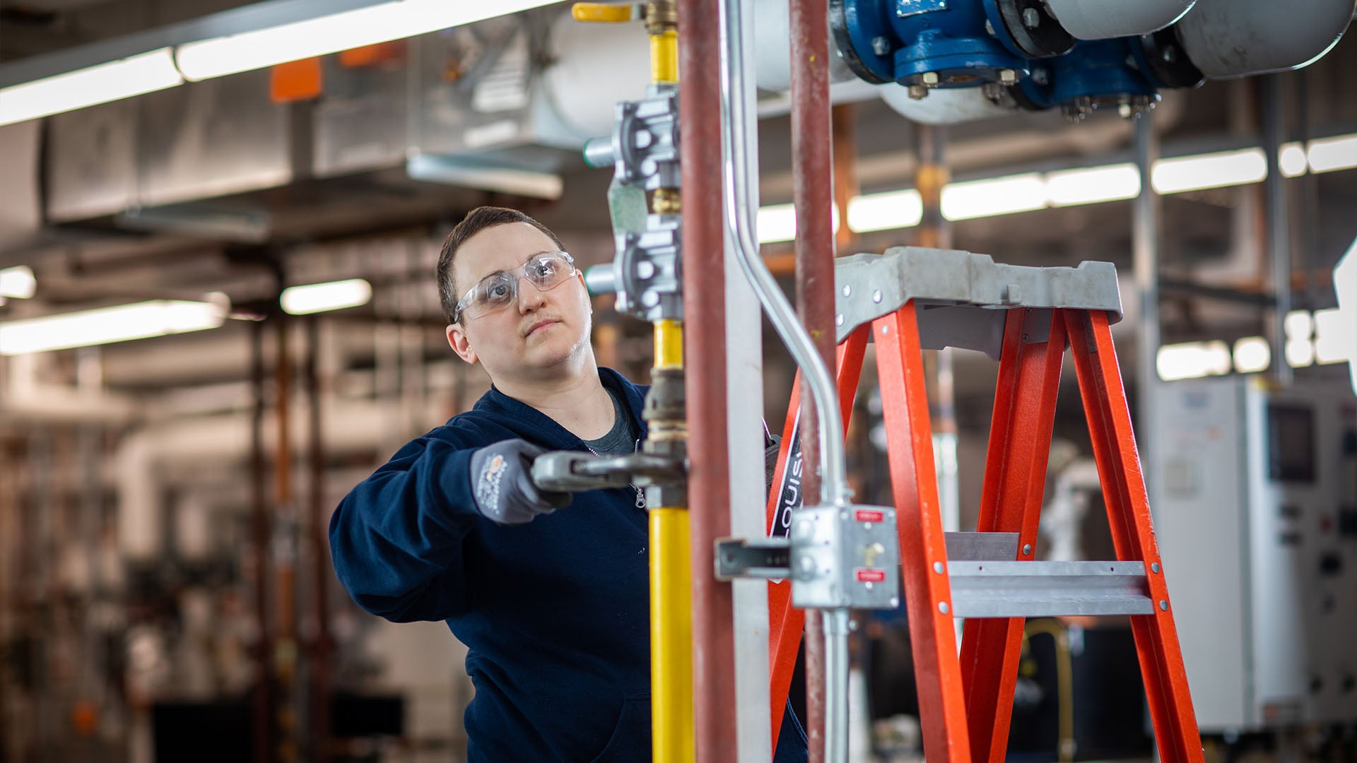Woman in safety glasses working with industrial pipes using a wrench, on a red a red ladder in a workshop setting.