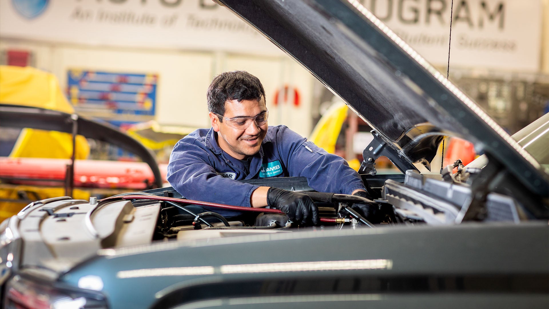 Man wearing safety glasses and gloves working under the hood of a car in an automotive workshop, with tools and vehicles in the background.