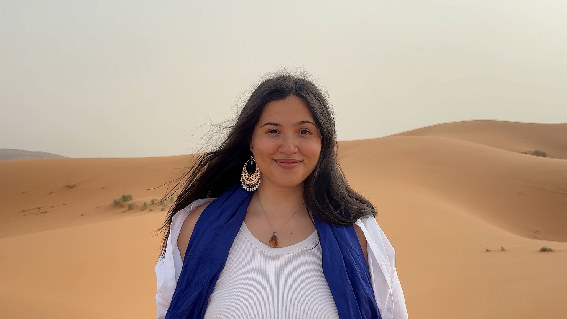 Person standing in a sandy desert landscape with rolling dunes, wearing a white top and blue scarf, with long dark hair and large earrings. Person standing in a sandy desert landscape with rolling dunes, wearing a white top and blue scarf, with long dark hair and large earrings.