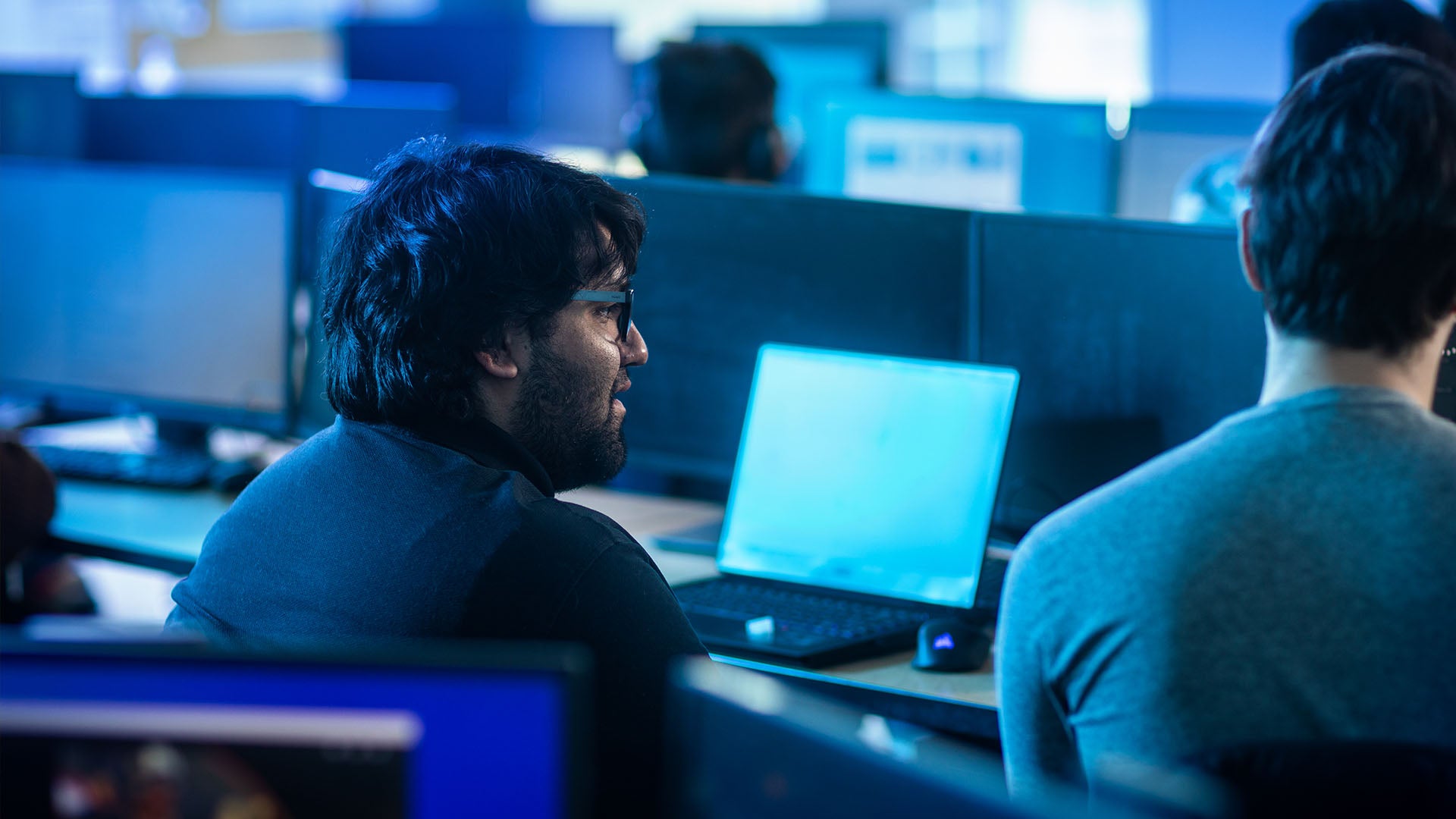 Two male students having a discussion in a computer lab with laptops open, surrounded by other students working on large monitors.