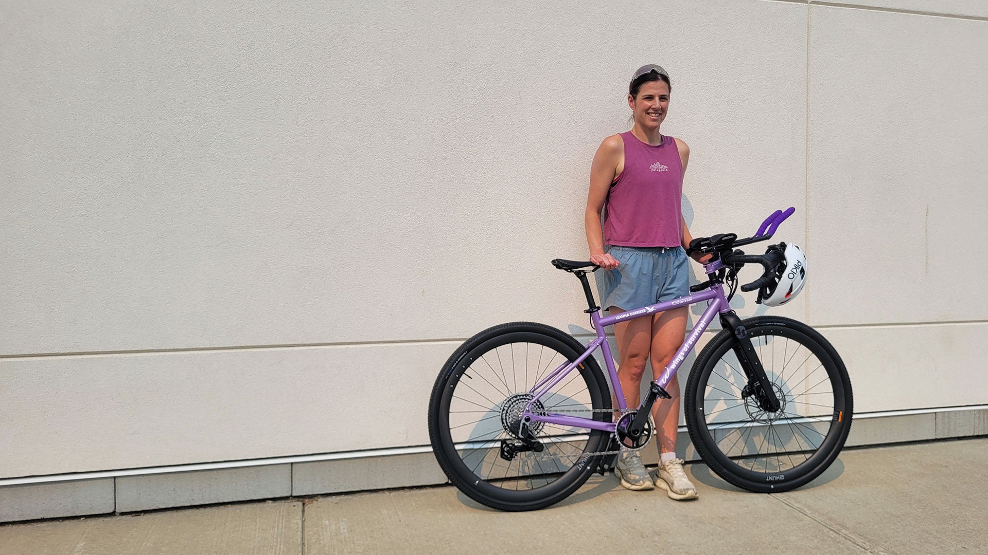 Person standing beside a purple bicycle and white helmet against a light-colored building wall. Person standing beside a purple bicycle and white helmet against a light-colored building wall.