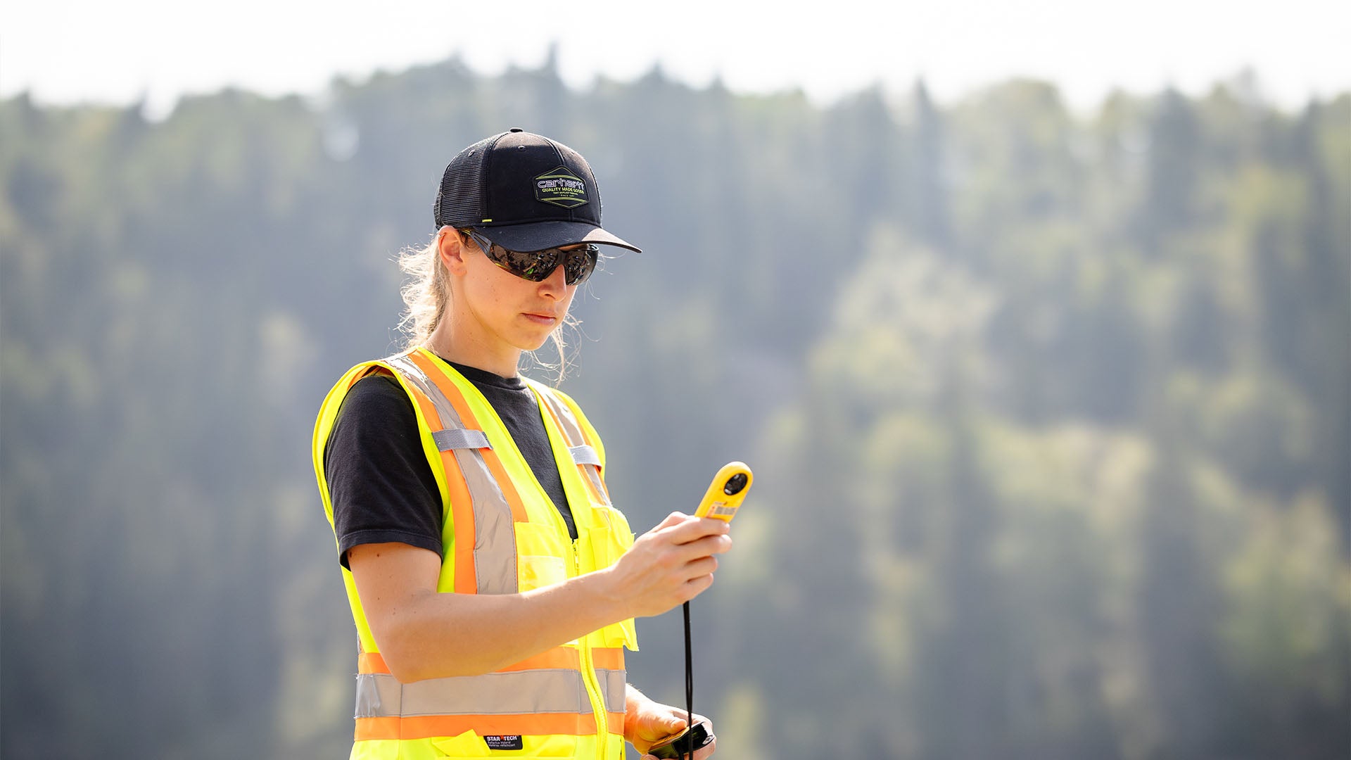 A female student wearing a high visibility vest, baseball cap and sunglasses looks at a weather meter.