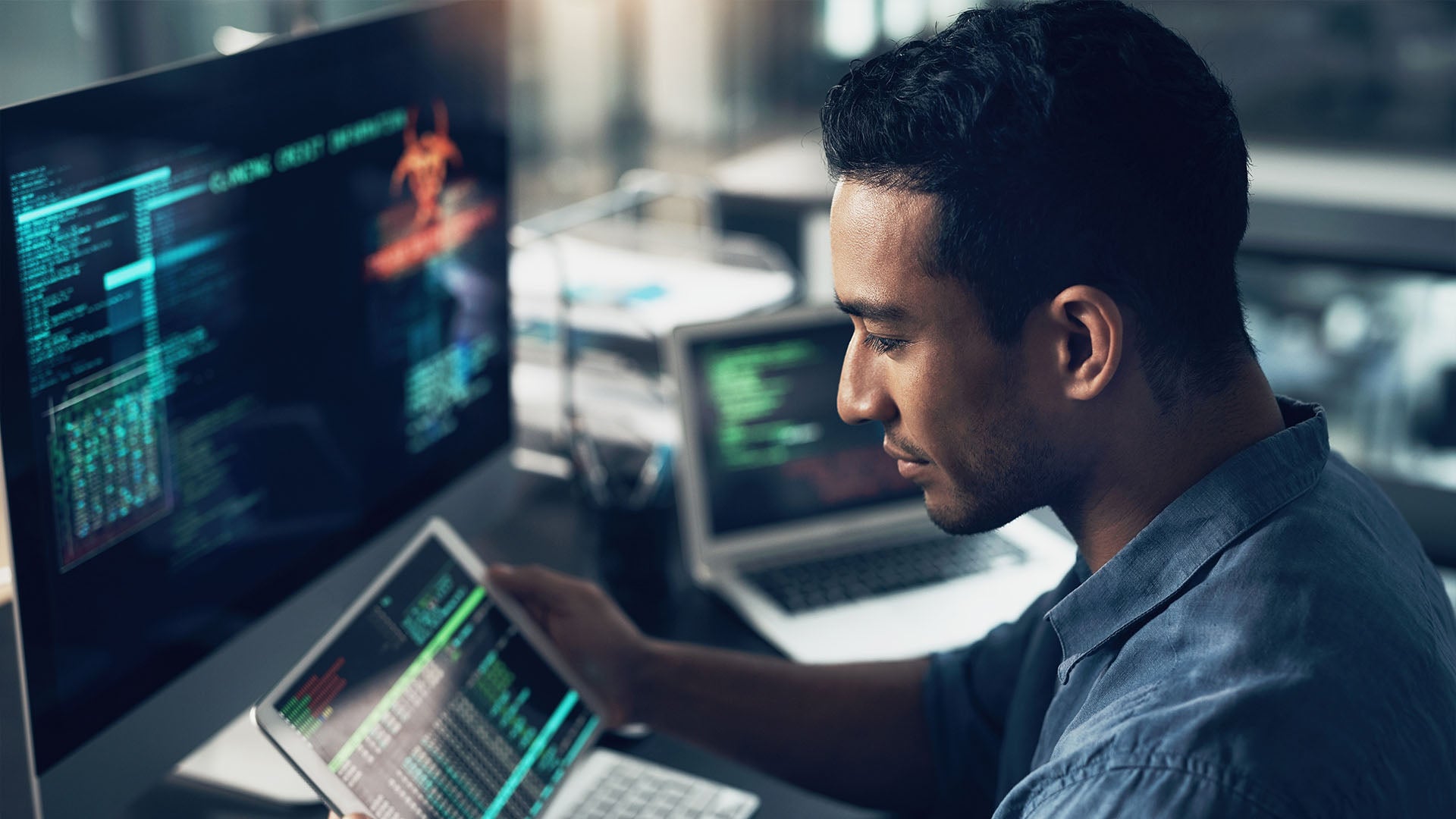 A focused professional analyzing data on a tablet while working on multiple monitors displaying code and cybersecurity-related visuals.