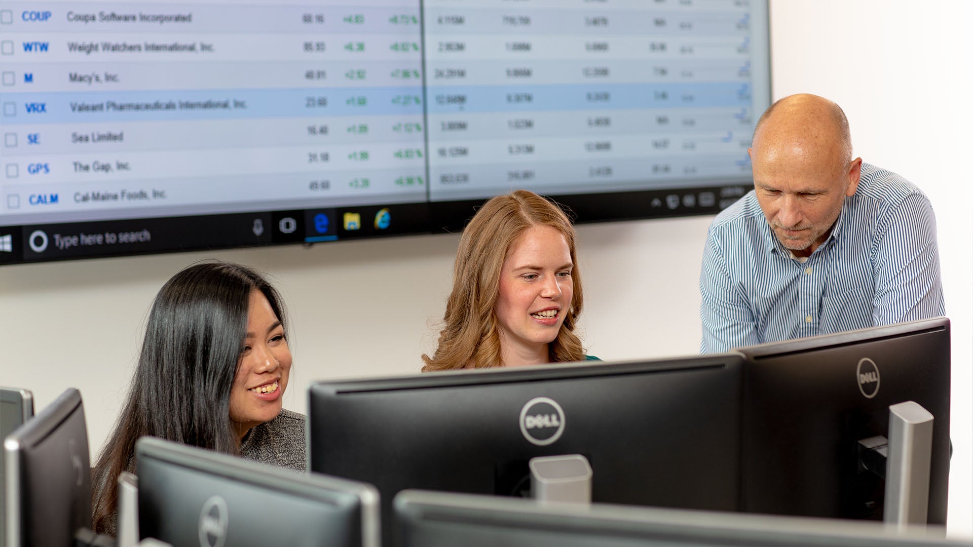 Two female students work at computers while an instructor observes. A projector screen in the background displays Yahoo Finance with stock market data.