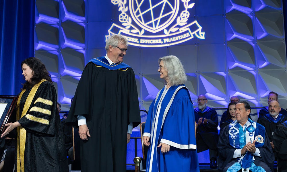 The image shows a formal ceremony taking place on a stage, likely at an academic or institutional event such as a convocation or award presentation.