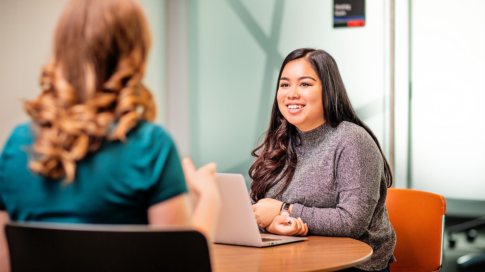 Two women having a professional conversation at a table, with one smiling and engaging attentively while using a laptop.