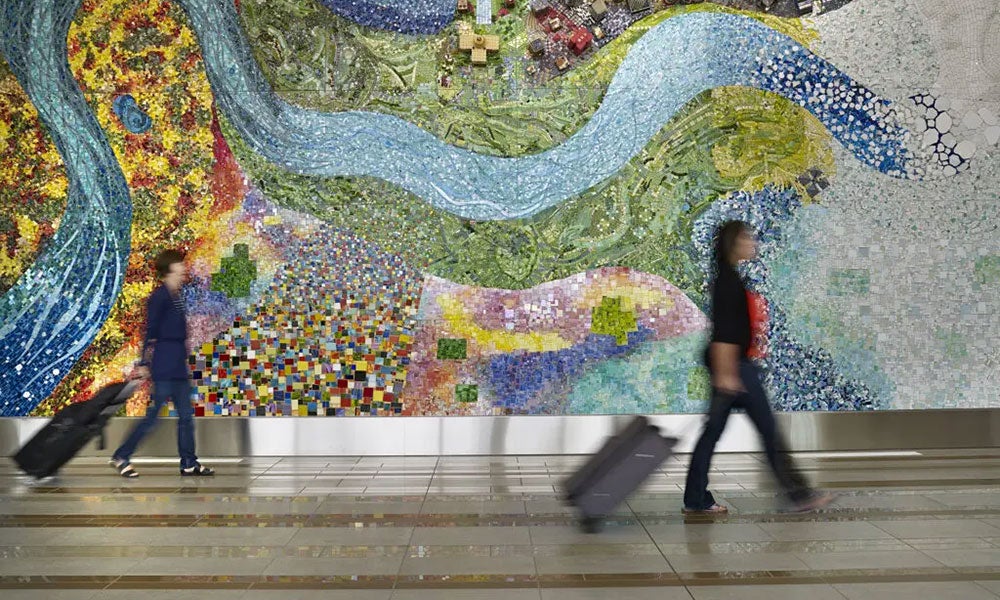 Travelers pulling rolling luggage walk through an airport concourse in front of a large, colourful mosaic mural featuring flowing river-like patterns and nature-inspired designs.