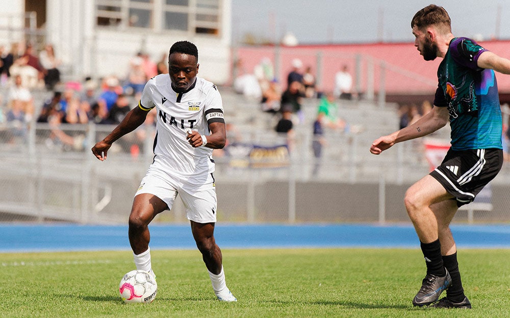 Men's soccer match on an outdoor field, with two players contesting the ball during active play in front of spectator seating and track facilities.