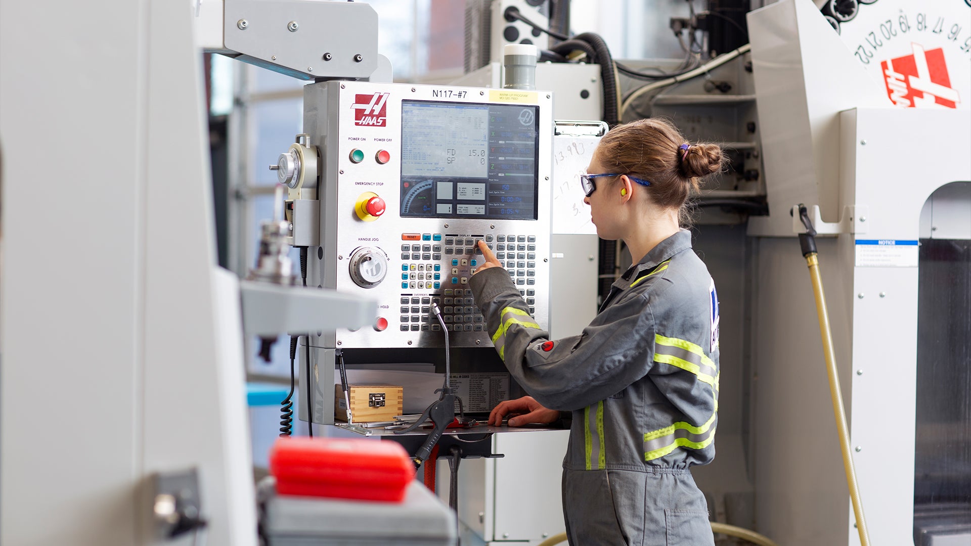 A woman wearing safety glasses and grey coveralls operates a Haas CNC machine.
