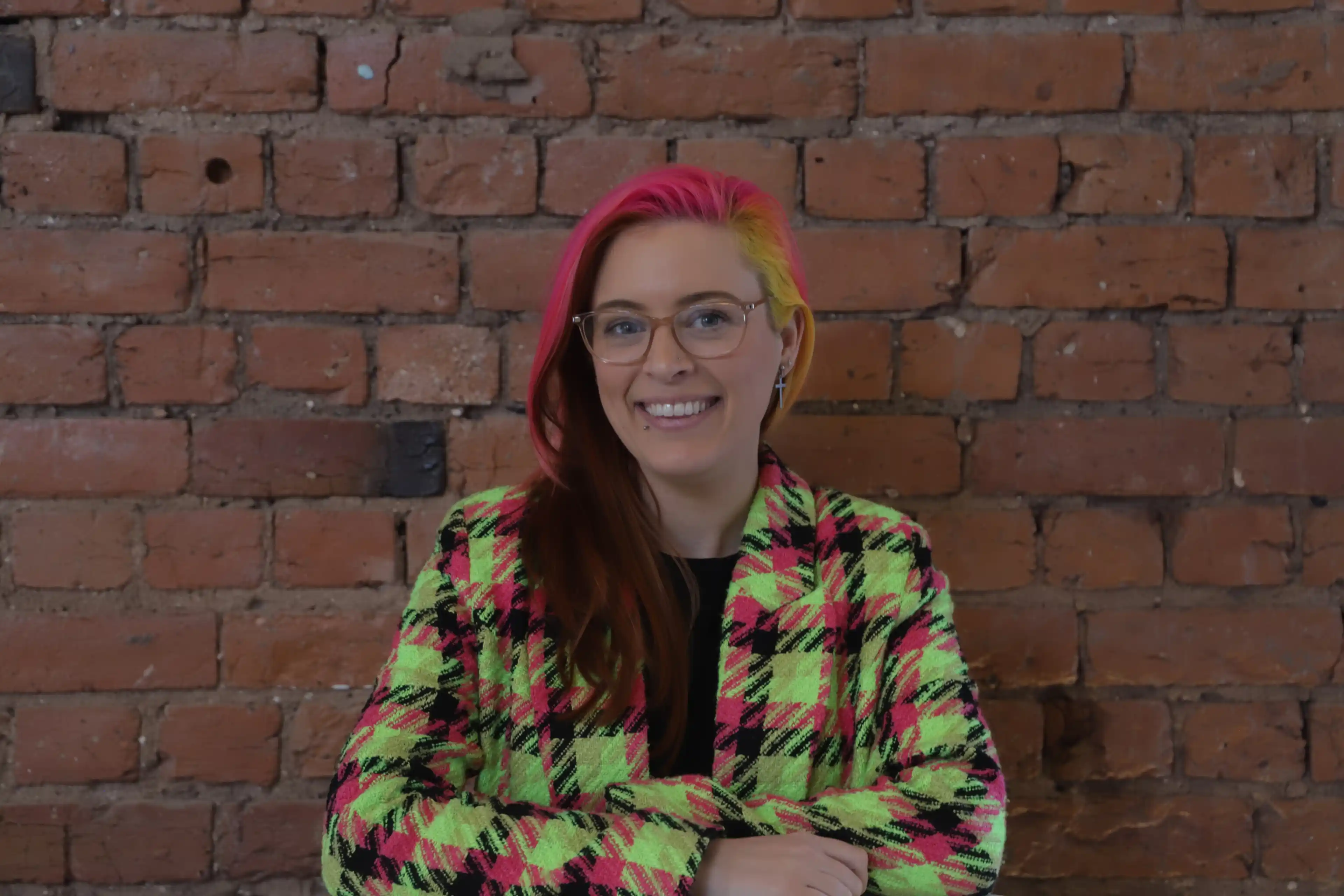 A woman wearing glasses with pink, yellow and dark red hair, wearing a yellow, pink and black jacket, standing in front of a redbrick wall