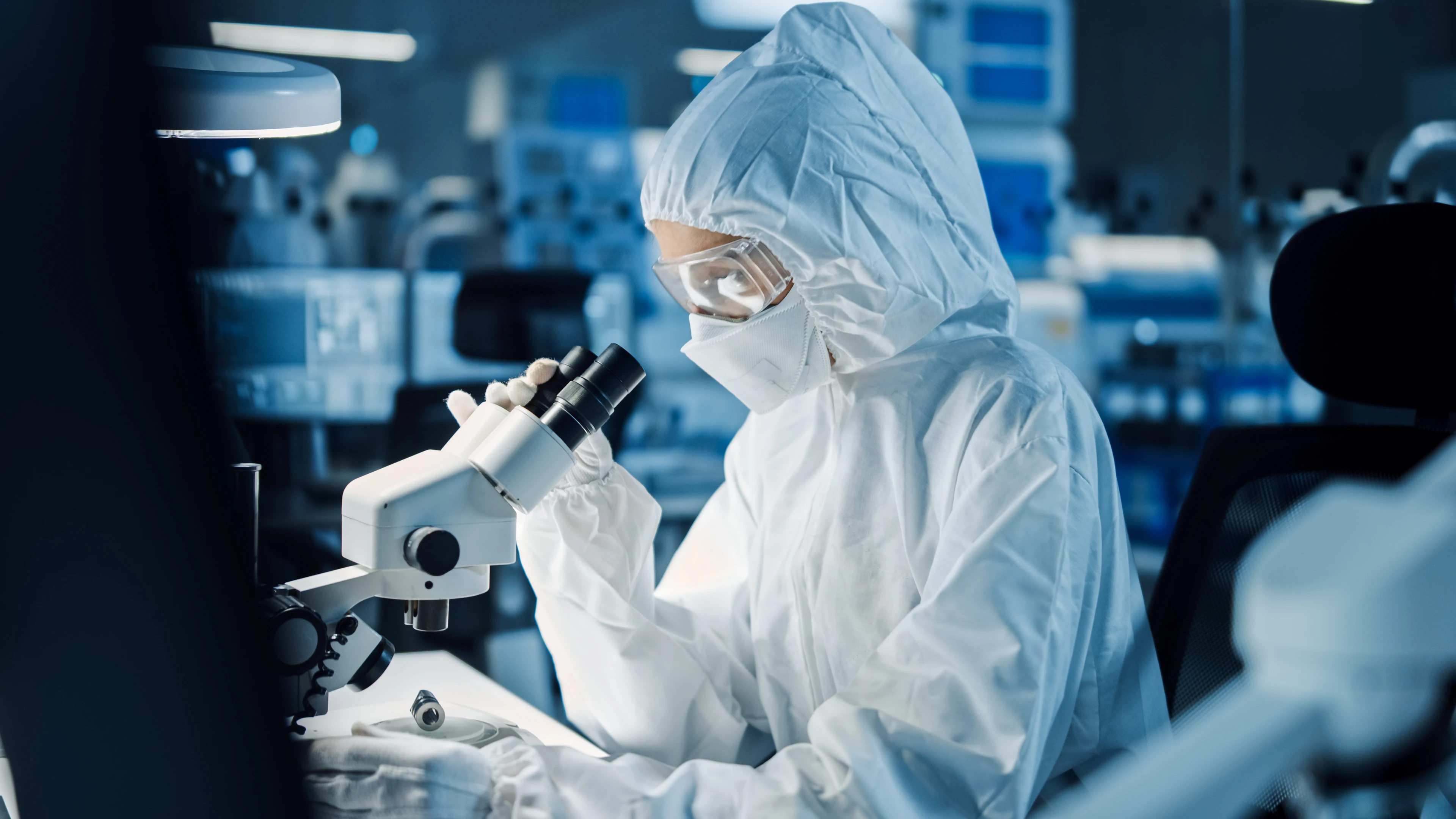 A laboratory worker wearing eye protection, mask, and full protective suit looks into a microscope.