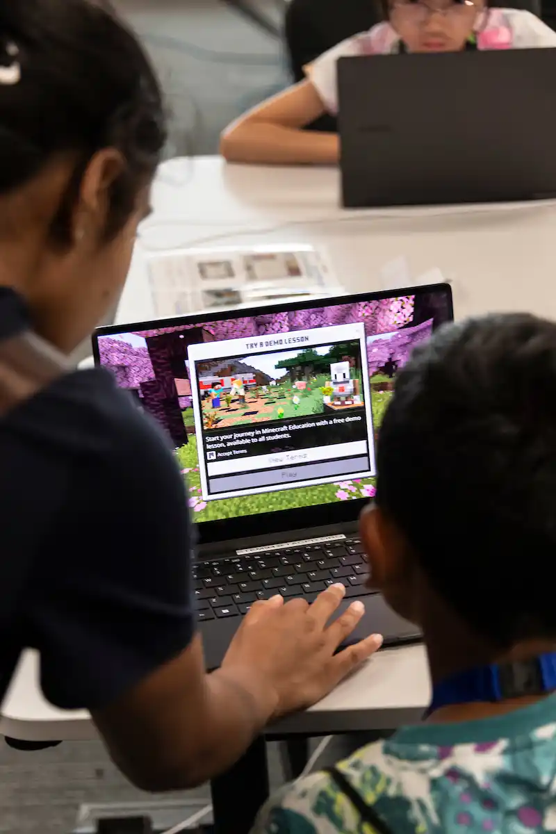 Teacher helping a child on a laptop who's playing the Minecraft led Skills Miner game by Enginuity