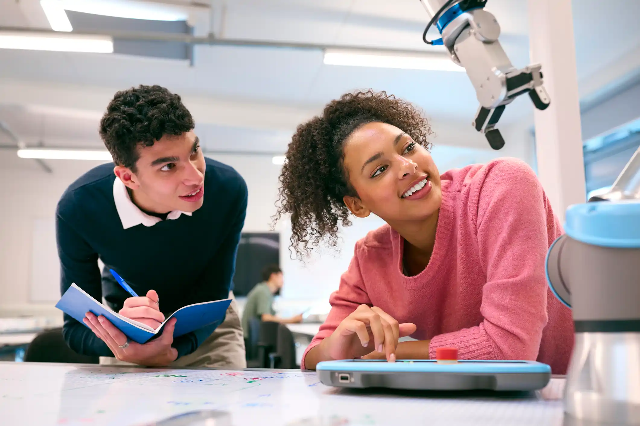 Two engineering student, male and female, looking at a robot arm and making notes in a classroom